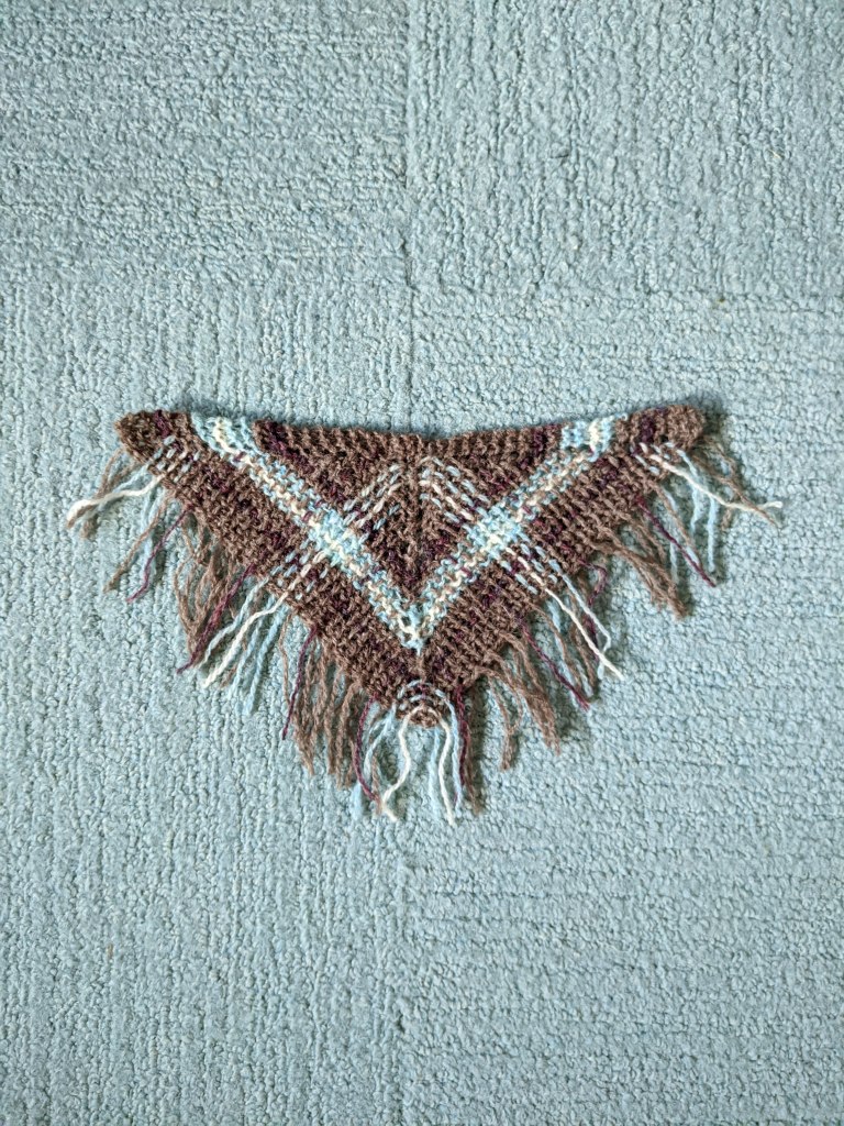 A small swatch for a plaid triangular shawl laid out on light blue carpet. The yarns are shades of medium brown, purple, white, and icy blue.